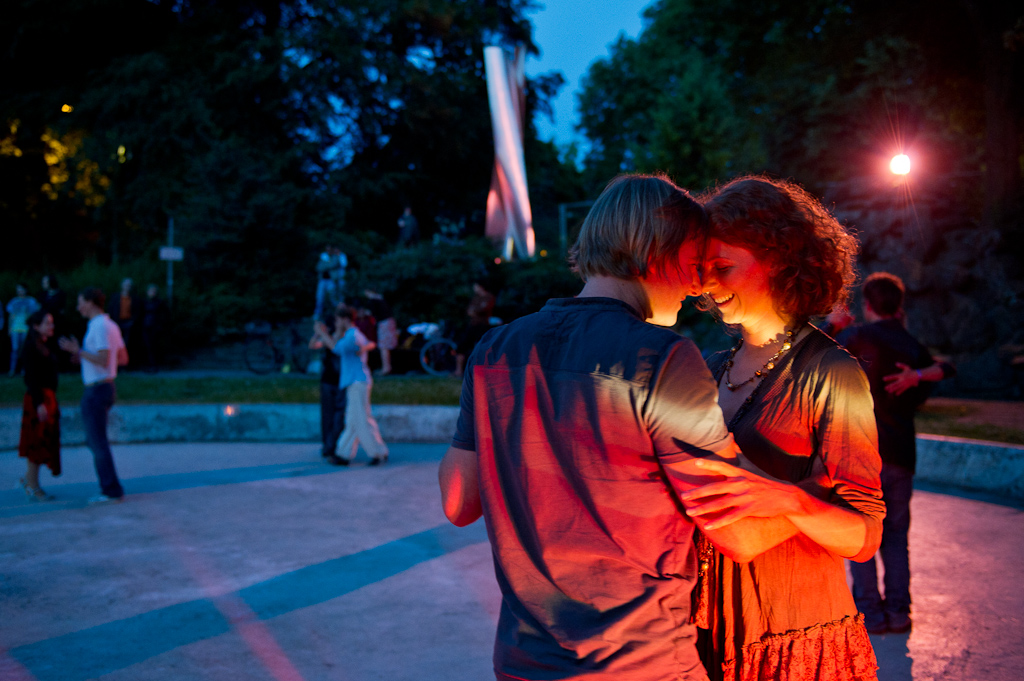 Ein Tanzpaar im Vordergrund tanzt in enger Umarmung, sie lächeln. Es ist Abend, ein rotes warmes Licht fällt auf sie, im Hintergrund ein Park und drei andere tanzende Paare. 